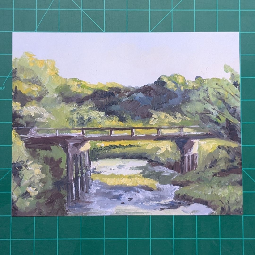 A landscape painting of a bridge over a stream, surrounded by green trees and foliage, displayed on a green cutting mat.